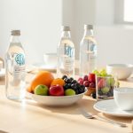 Table lumineuse de petit-déjeuner avec trois bouteilles d’eau minérale en verre, des fruits frais et un verre transparent dans une ambiance minimaliste et apaisante.