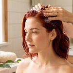 Femme aux cheveux auburn fraîchement colorés appliquant doucement un soin botanique apaisant sur son cuir chevelu dans une salle de bain lumineuse, avec des accessoires de routine capillaire vegan.